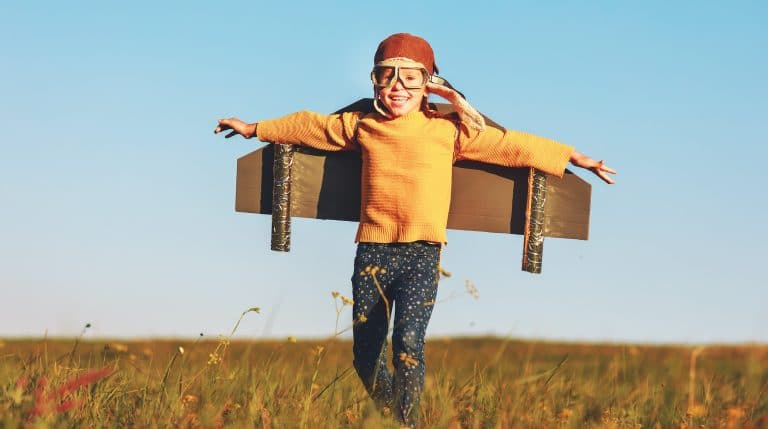 A child, basking in the joy of visitation, wears a pilot's cap and goggles while smiling and running through a grassy field. Attached to their back is a homemade cardboard airplane. The sky is clear and blue.