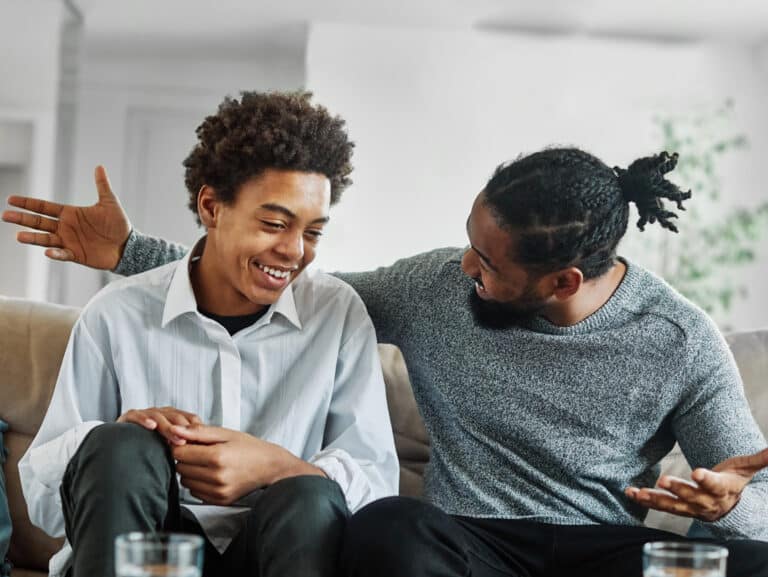 A man with a beard and hair in a bun is sitting on a couch, smiling and gesturing as he talks to a younger man next to him, who is also smiling. The relaxed living room setting reflects their effort in communicating with kids for better relationships.